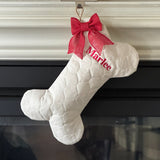 A bone-shaped light beige quilted Christmas stocking with a red bow and red embroidered name 'Marlee', hanging in front of a fireplace.