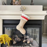 BurlapBabe burlap Christmas stocking with ivory fleece cuff, red ribbon trim, and two wooden buttons on a fireplace.